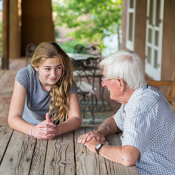 Girl having a conversation with an elderly man while sitting at a picnic table outside on a covered portch area.